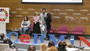 En el escenario: una joven en silla de ruedas, una mujer de pie, y otro hombre de pie, hablando. Una mujer le sujeta un micrófono.