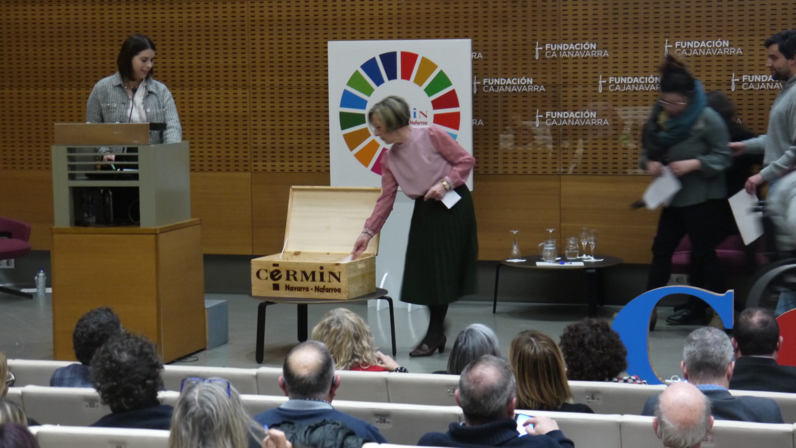 Una de las mujeres que ha leído el Manifiesto mete una tarjeta en una caja de madera. Detrás hay un cartel con el logo de CERMIN y de los ODS.