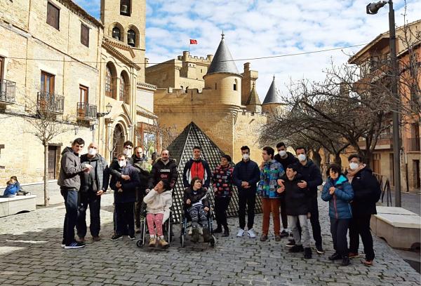 Fotografía de un grupo de personas frente al castillo de Olite. Llevan abrigos, dos de ellas están en silla de ruedas y varias llevan mascarillas.