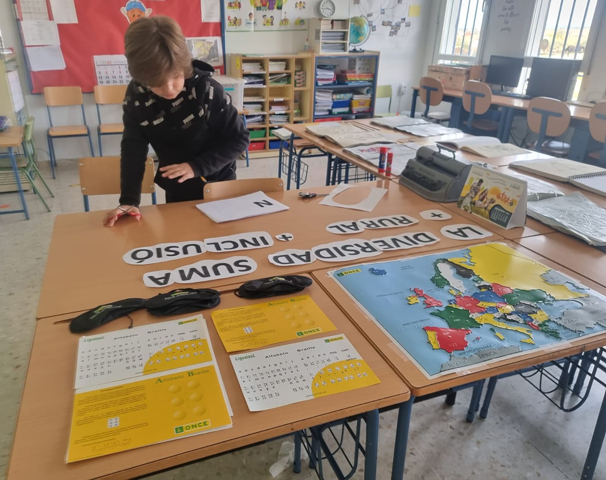 Joven escolar en un aula, trabajando con material didáctico de ONCE: hay un mapa de Europa y láminas de alfabeto braille. También está trabajando con un eslogan que dice "La diversidad suma". En otros papeles aparecen escritas las palabras "rural" e "inclusión".