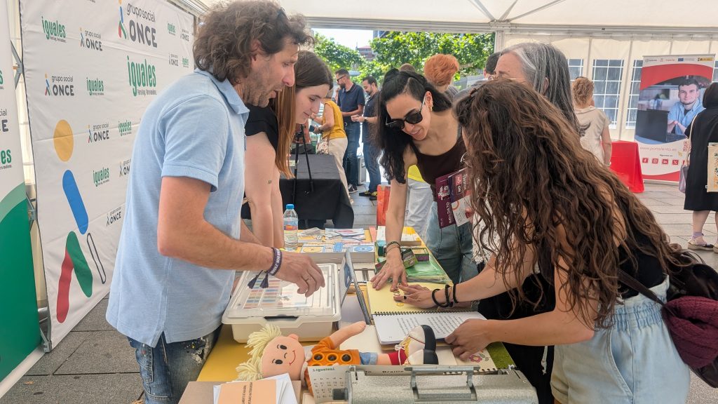 Espacio informativo de Braille en la carpa de grupo social ONCE Navarra. Varias personas están alrededor de una mesa viendo y tocando material para el aprendizaje del braille.
