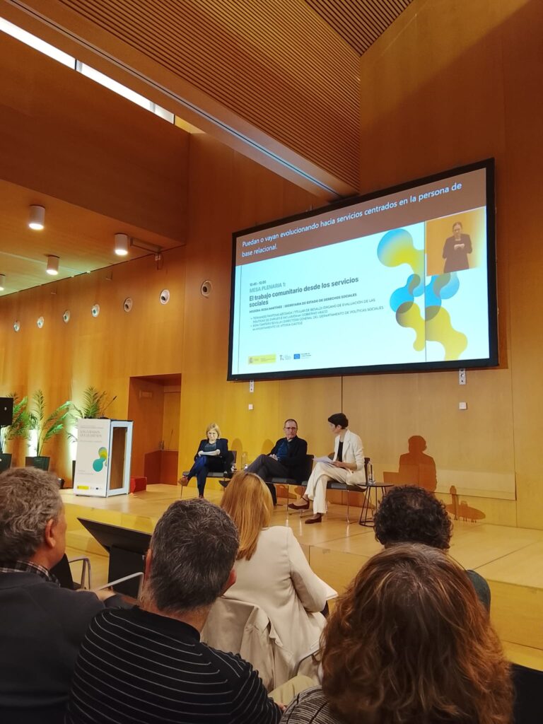 momento de una ponencia, tres personas en el escenario. Detras la pantalla de la jornada